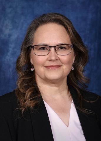 Formal professional headshot of an individual with shoulder-length brown hair wearing glasses, a dark blazer, and a light-colored blouse, facing forward against a softly textured blue studio background.