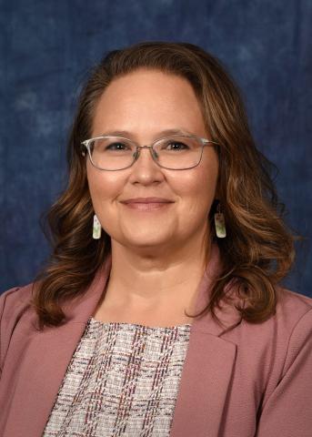Formal professional headshot of an individual with shoulder-length brown hair wearing glasses, a pink blazer, and a plaid blouse, facing forward against a softly textured blue studio background.
