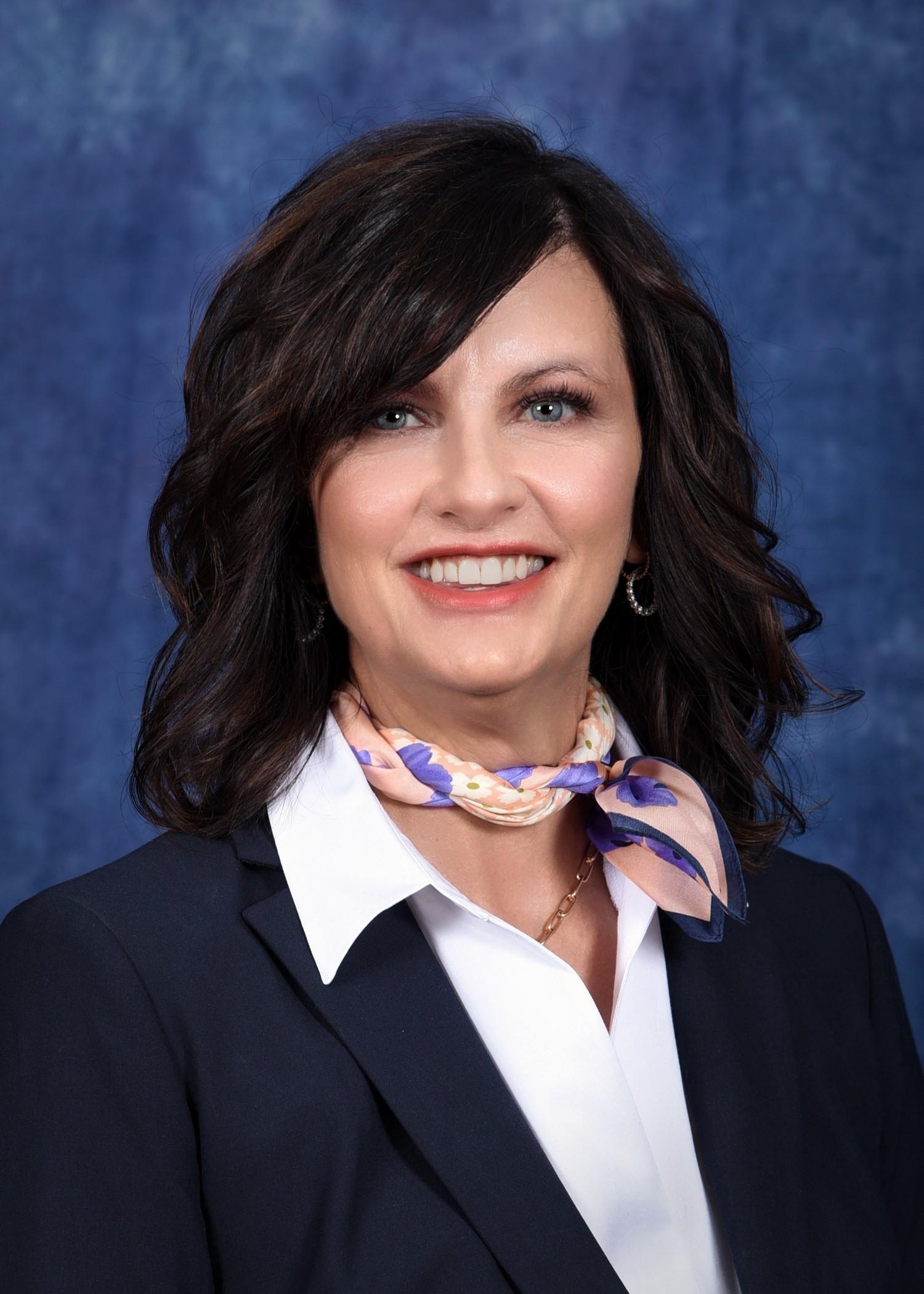 Formal professional headshot of Lori Barber, President of the College of Eastern Idaho. The individual has shoulder-length dark brown hair and is wearing a dark blazer over a white collared shirt with a light patterned scarf tied at the neck and small hoop earrings. The portrait is set against a softly textured blue studio background.
